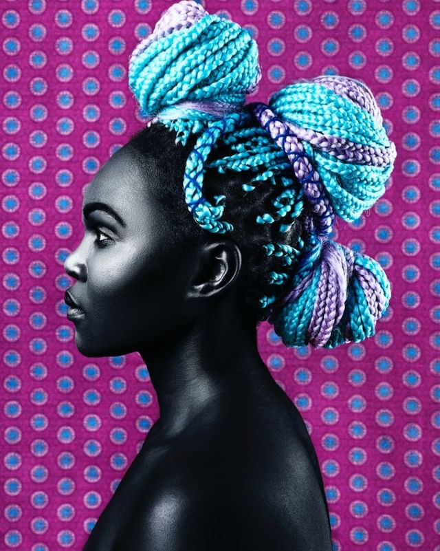 Profile of a Black woman with blue and purple braided hair styled in buns, against a purple polka dot background.