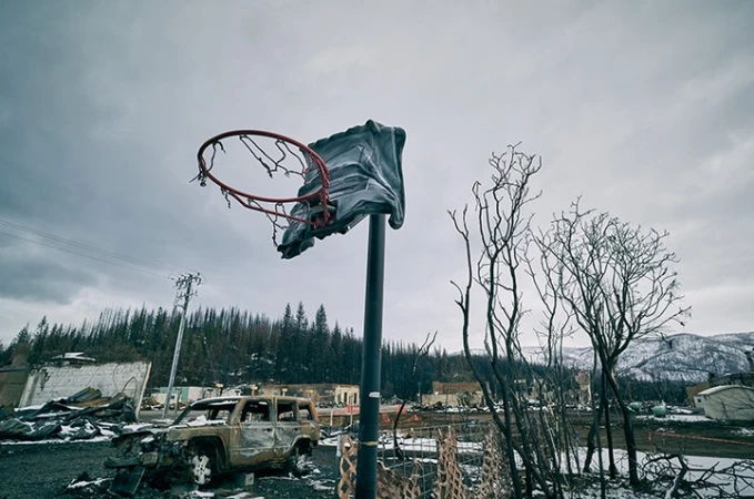 Damaged basketball hoop stands amid destroyed buildings and vehicle wreckage under a cloudy sky, conveying post-disaster aftermath.