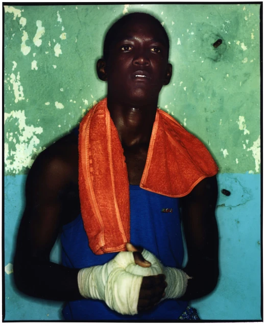 A close-up of a male boxer of African descent with wrapped hands and an orange towel around his neck, posing indoors against a blue-green wall.