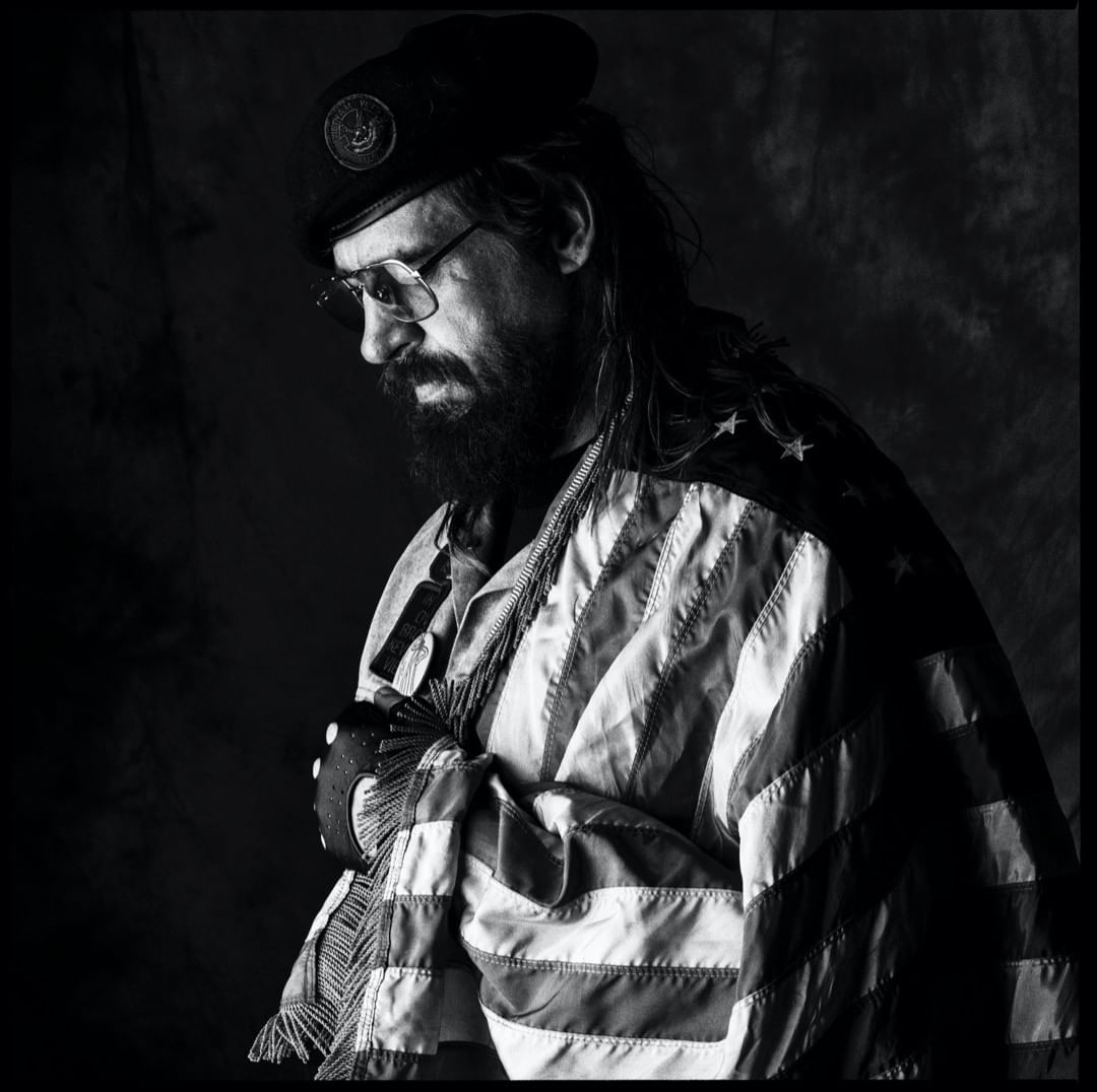 Monochrome portrait of an adult male with a beard wearing glasses, beret, and a US flag draped over his shoulders in dark setting.