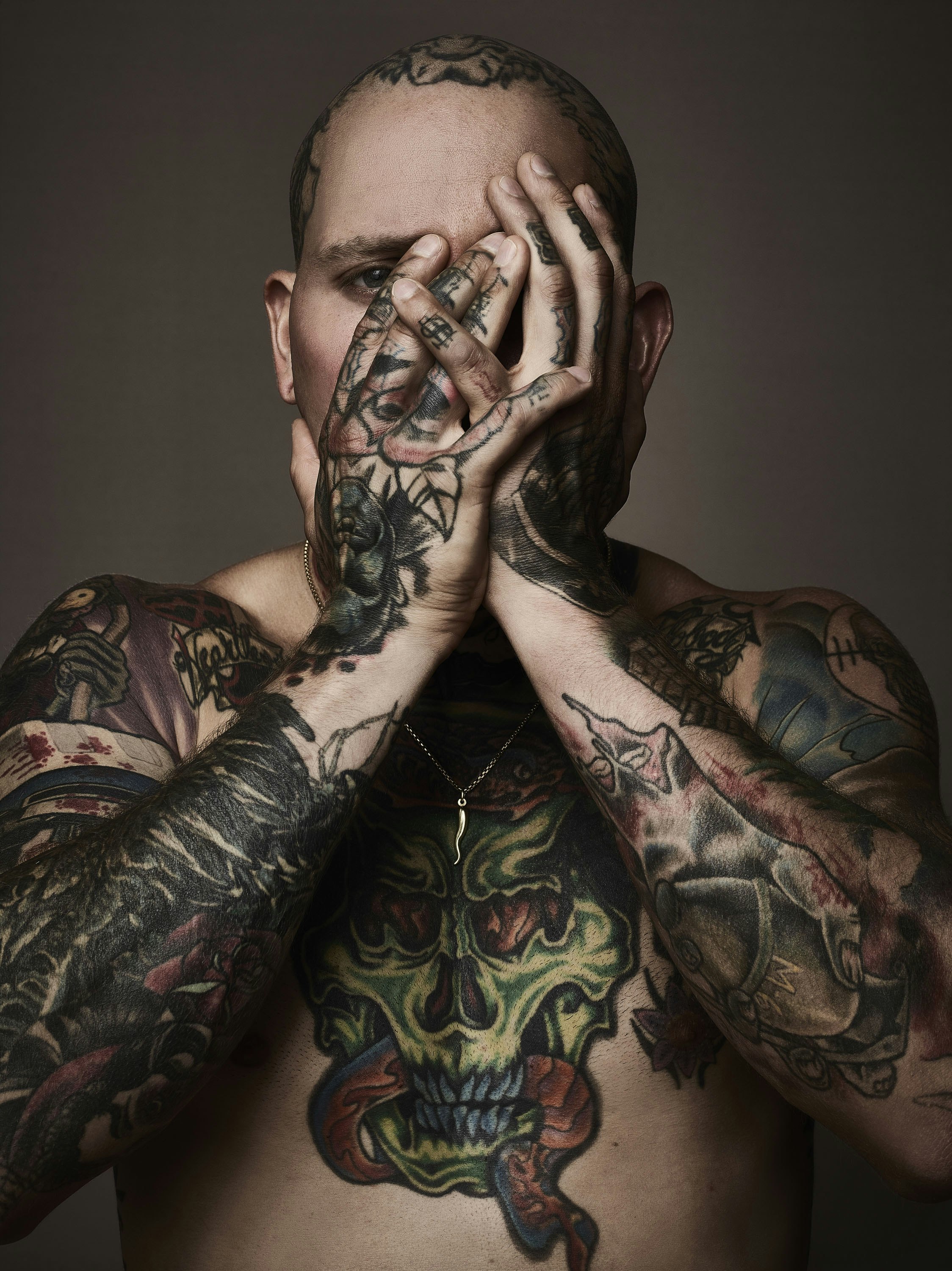 Portrait of an adult male with extensive tattoos covering his body and face, obscuring his face with his hands in a studio setting.