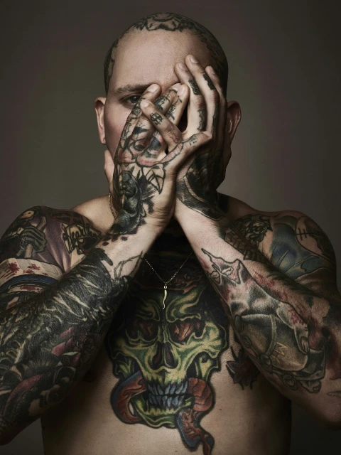 Portrait of an adult male with extensive tattoos covering his body and face, obscuring his face with his hands in a studio setting.