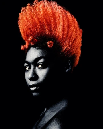 Portrait of a Black woman with voluminous bright orange hair and small Bantu knots against a black background.