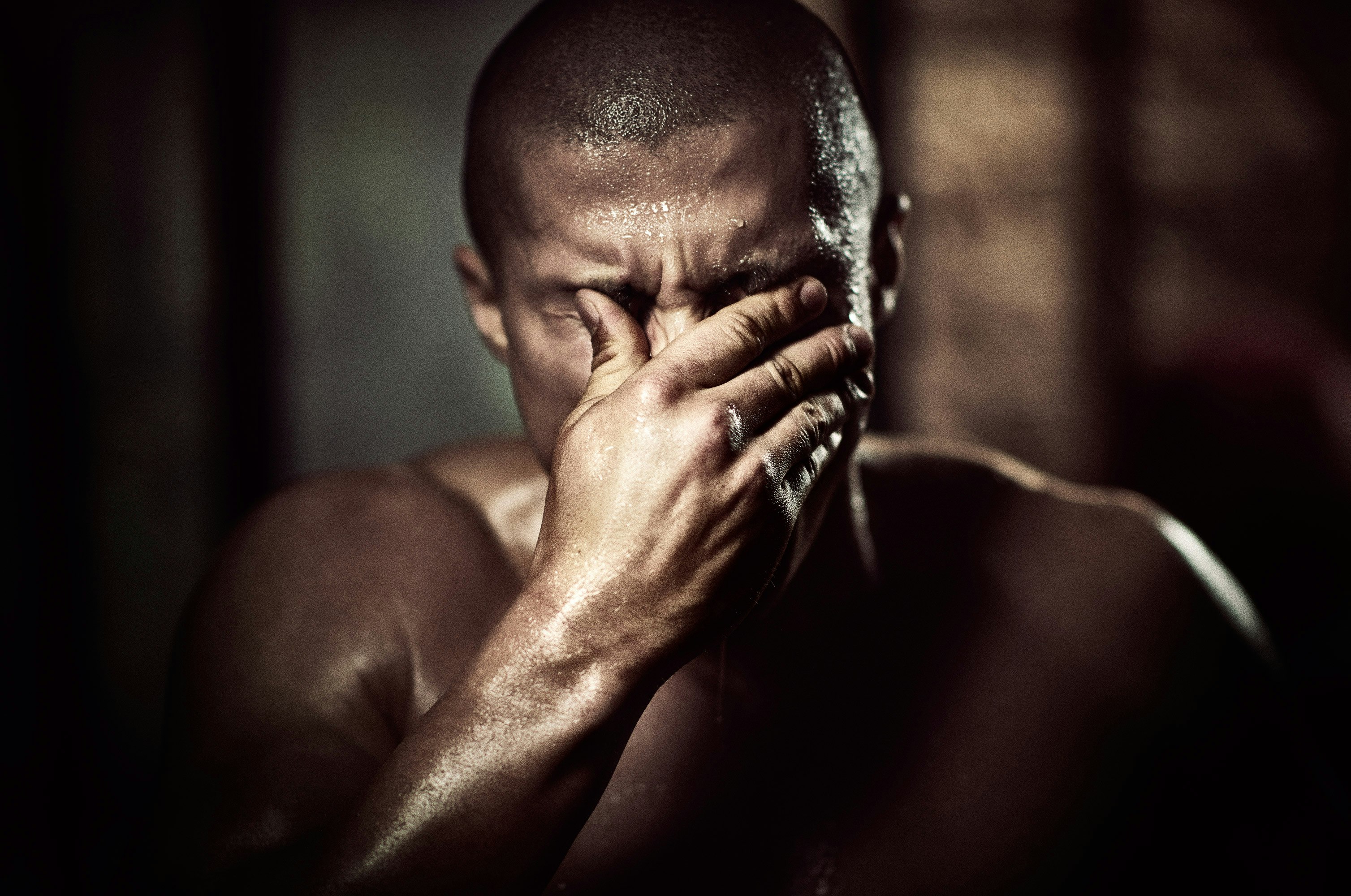 Sweaty, bald man in dark environment covering his eyes with hand. Close-up shot of adult male with intense expression in low light.