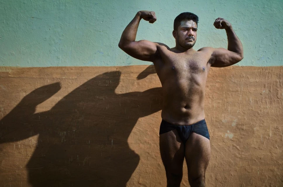 Man posing with flexed biceps in front of a textured blue and brown wall, casting a shadow to the left; wearing minimal black briefs.