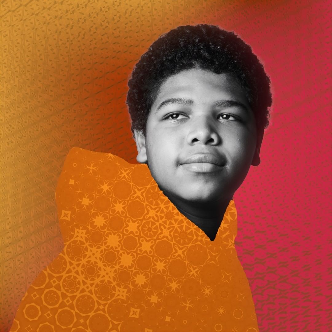Black and white portrait of a boy with an afro, wearing an orange hooded sweatshirt with a patterned design over a colorful background.