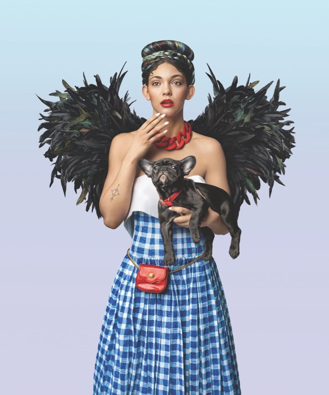 Woman in Dorothy costume holds a black French bulldog with feather wings against a gradient blue background in a studio shot.
