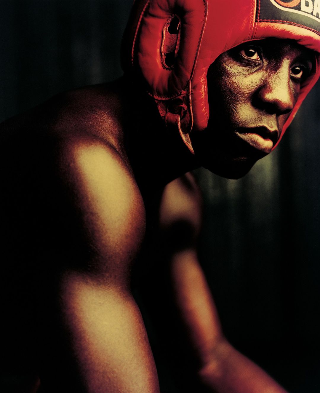 Portrait of a muscular adult male boxer with dark skin wearing a red boxing headgear in a dimly lit, close-up studio shot.