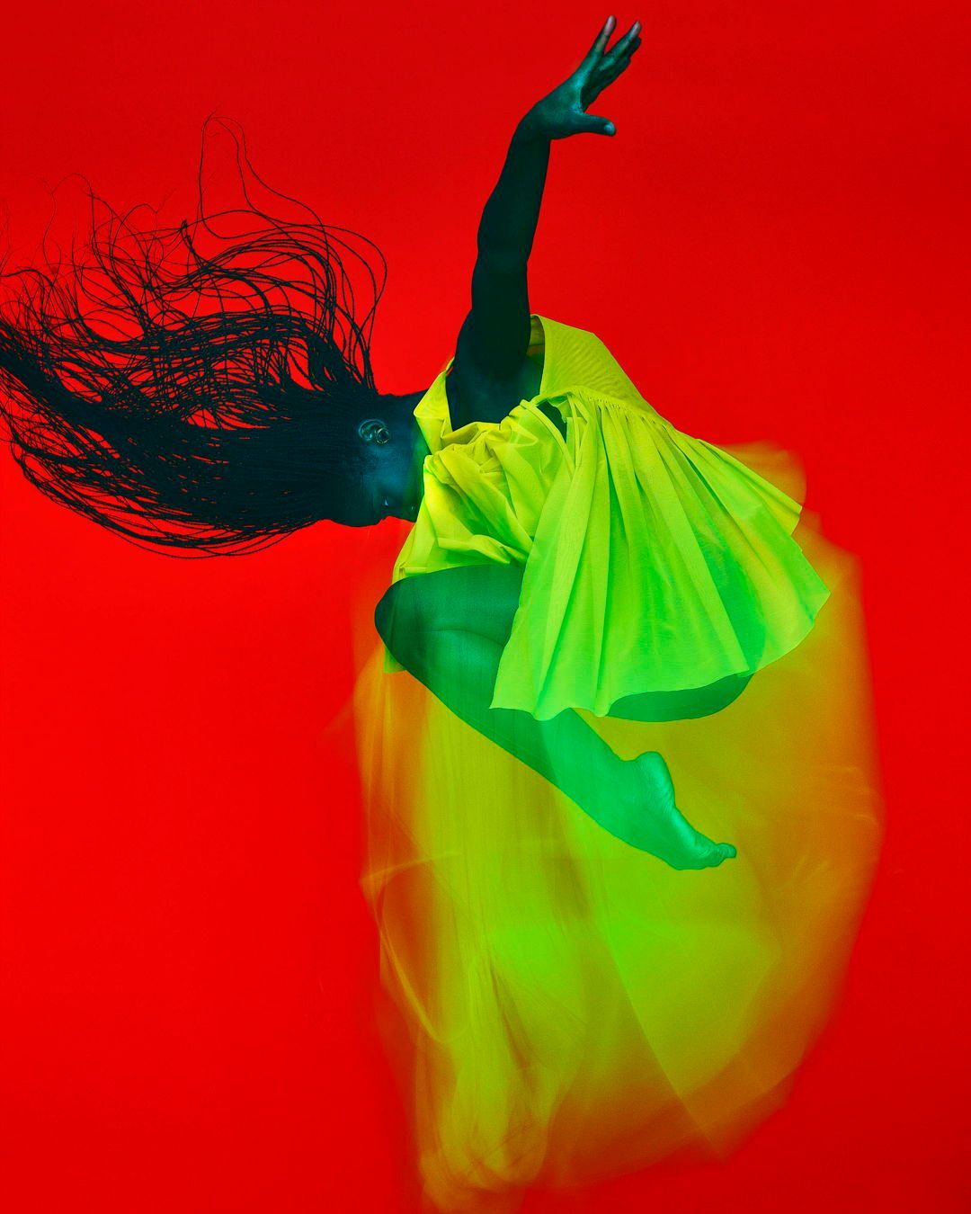 Woman in motion wearing a bright yellow dress against a red background, creating a dynamic and striking visual. Long dark hair flowing.