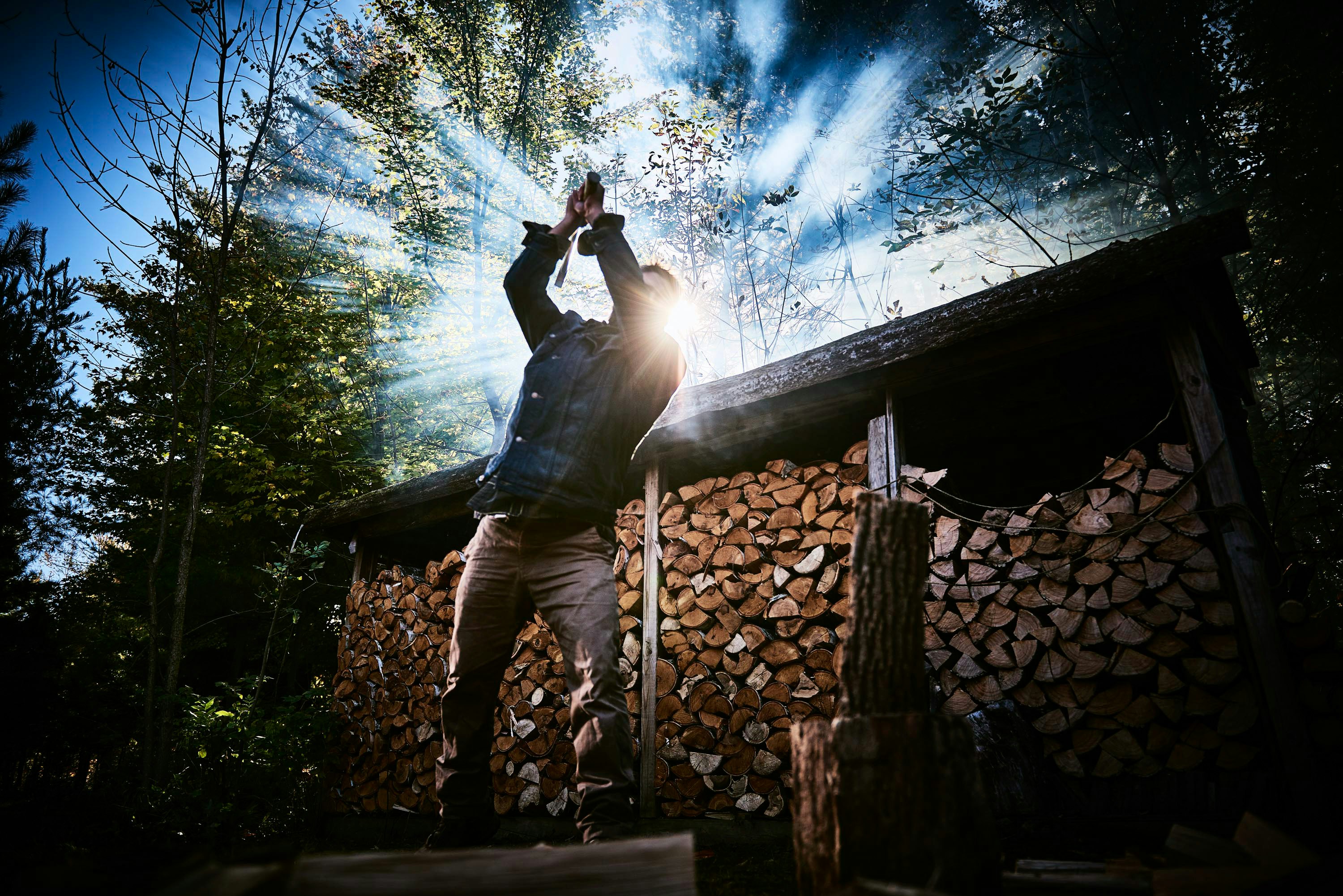 Man chopping wood with an axe in a forest clearing near a wood pile, sunbeams shining through the trees, low angle.