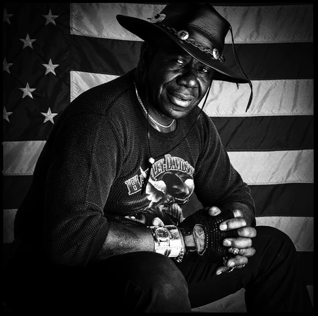 Black and white portrait of African American man in cowboy hat with American flag background. Man is seated, wearing a Harley Davidson shirt.