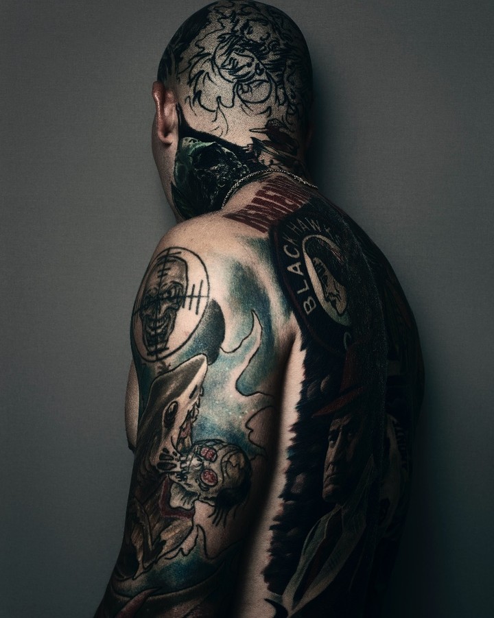 Portrait of an adult male with extensive head and back tattoos featuring a black and white color palette in an indoor setting.