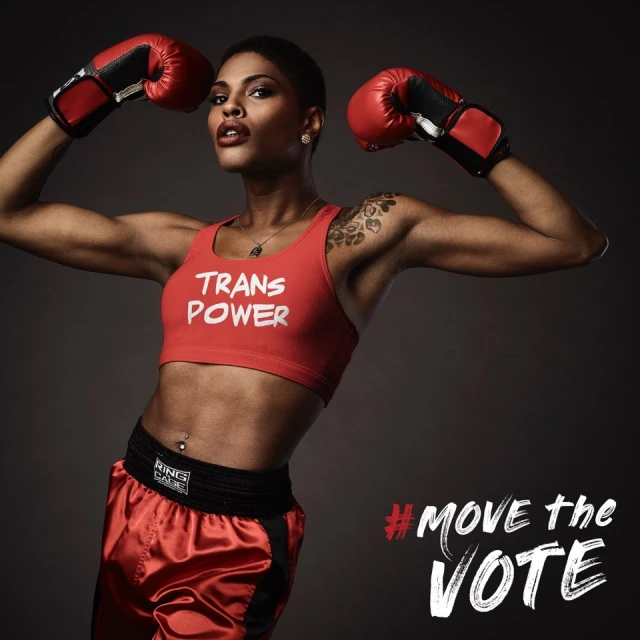 Athletic woman flexing biceps while wearing boxing gloves and 'Trans Power' shirt in a fitness campaign against a grey background.