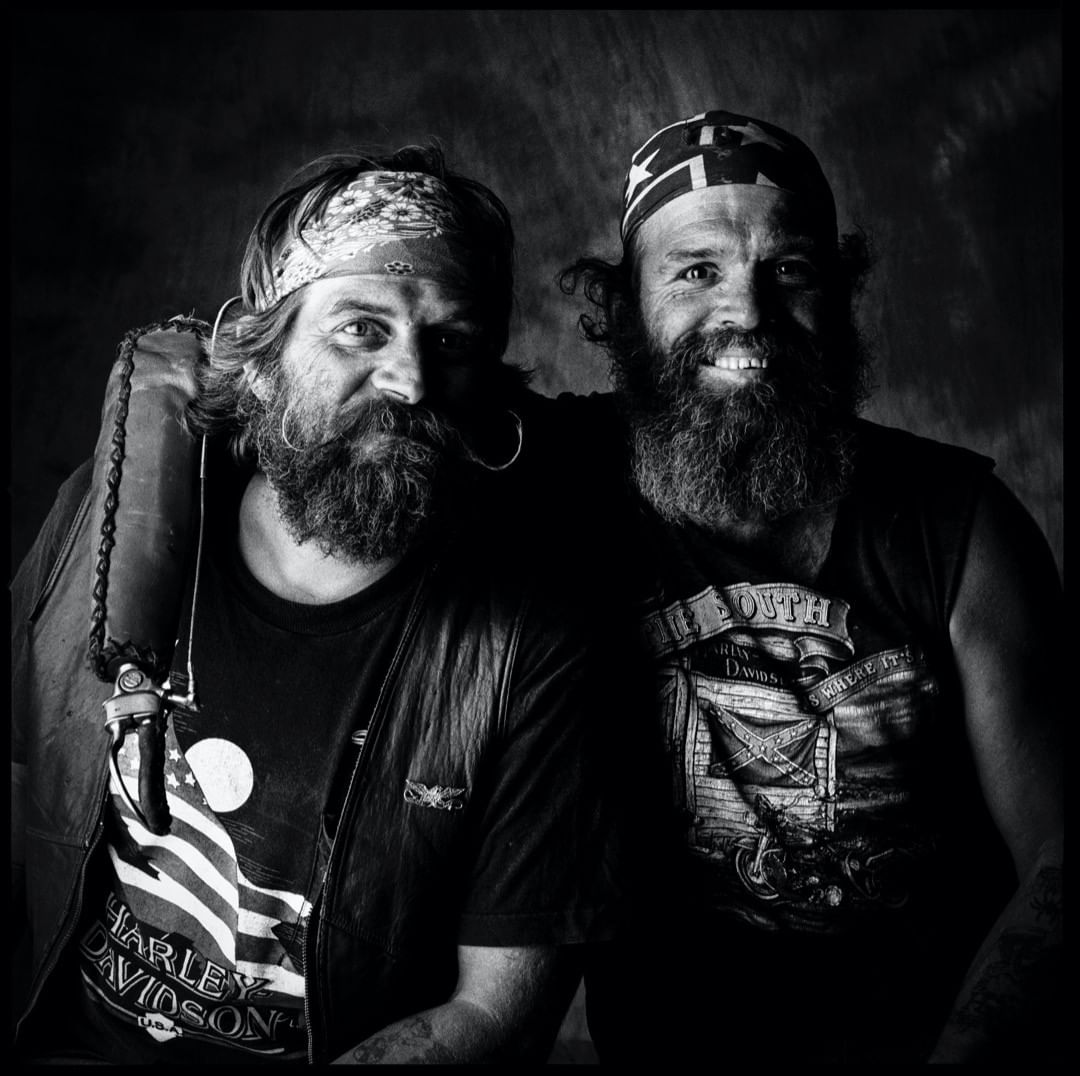 Two men with beards pose indoors, wearing bandanas and t-shirts with American flag and motorcycle imagery, in a black and white portrait.