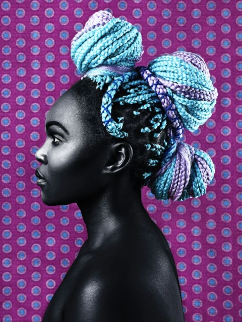Profile portrait of a young Black woman with braided turquoise and purple hair styled in buns against a purple polka dot backdrop.