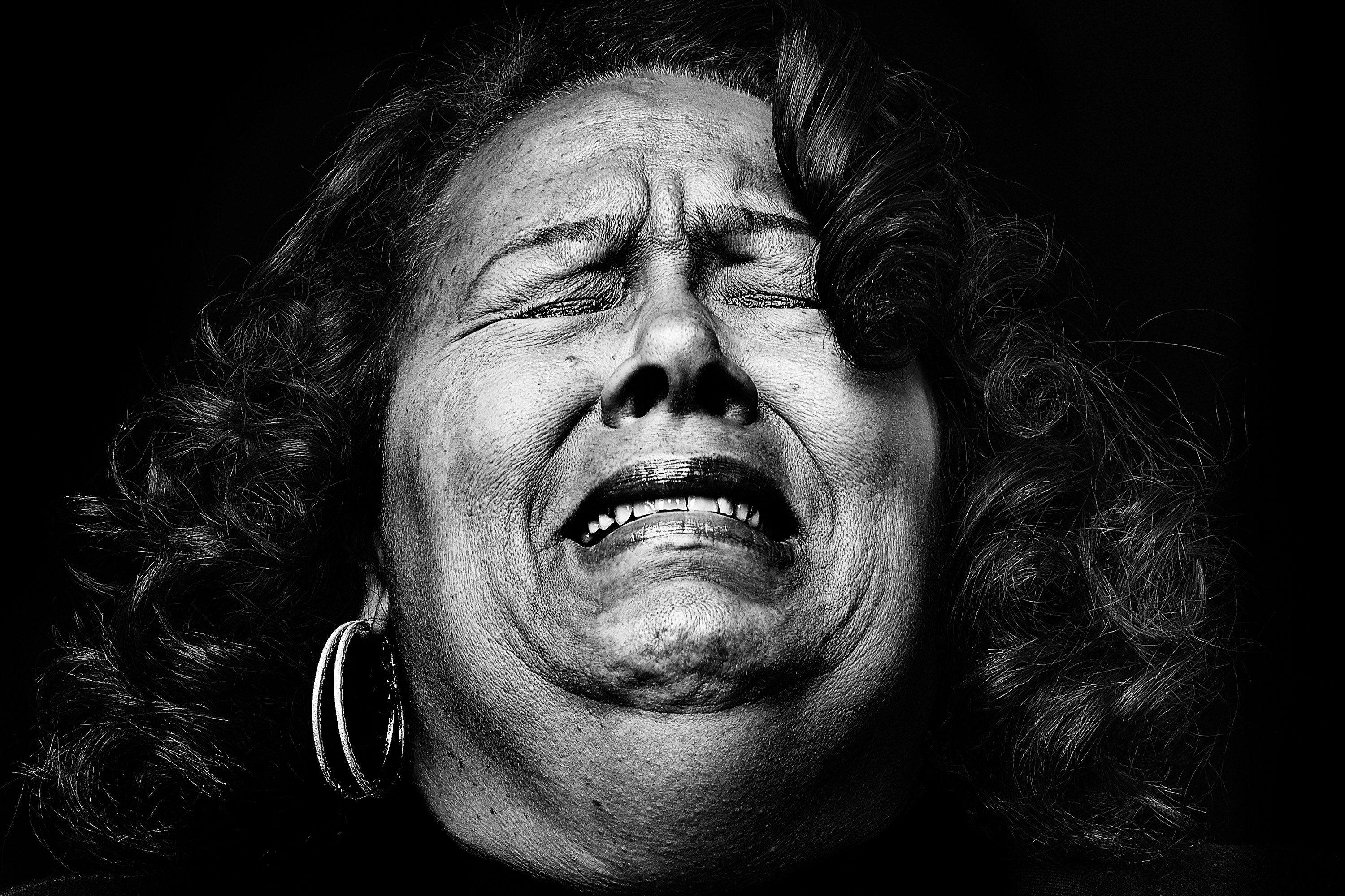 Close-up black and white portrait of a mature woman with curly hair, closed eyes, and a distressed expression in front of a dark background.