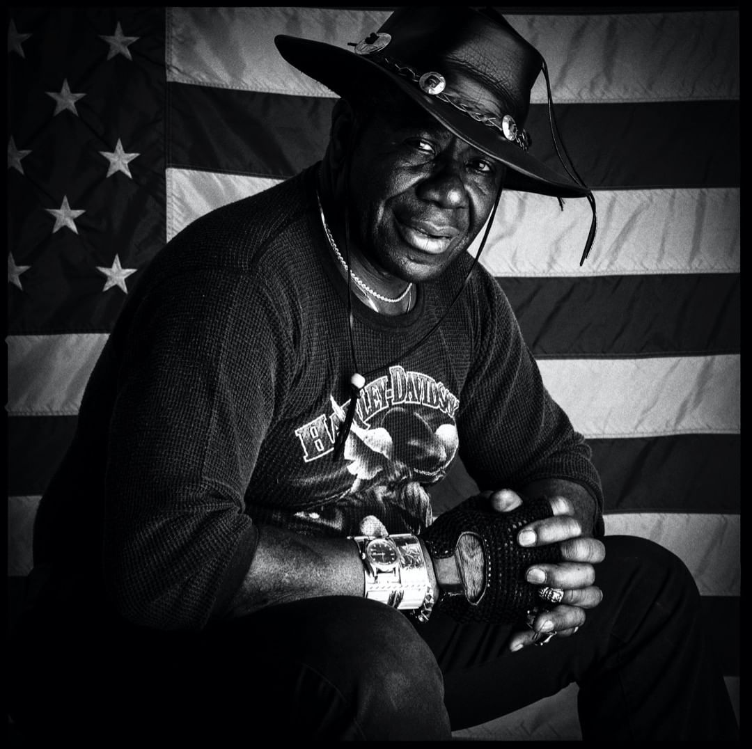 Monochrome portrait of an African American man wearing a cowboy hat and patterned shirt against a striped American flag backdrop.