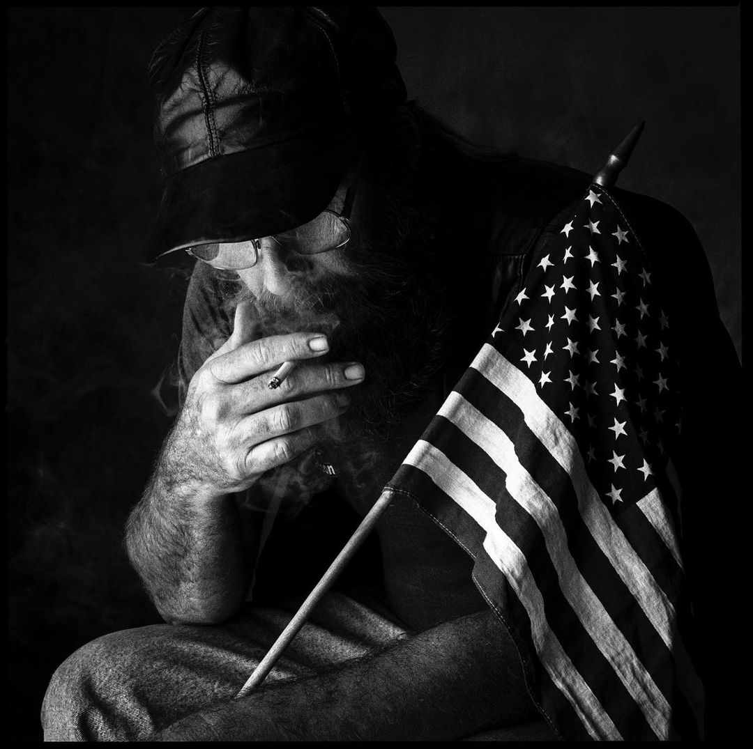 Black and white portrait of a man with long beard and hat, smoking a cigarette and holding an American flag against a dark background.