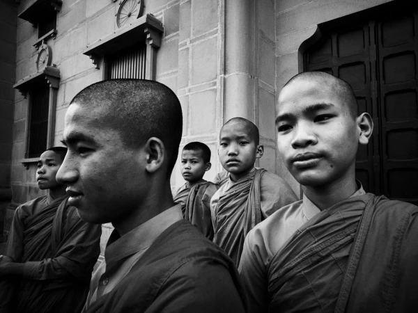 Black and white image of a group of young Buddhist monks in robes standing outside a stone building with windows and doors.