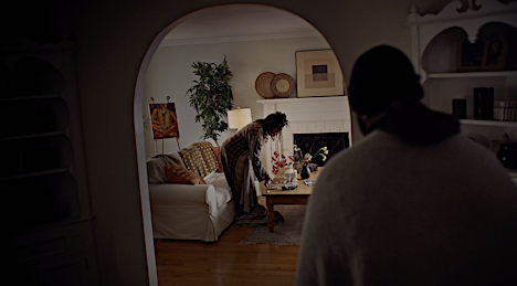 Indoor domestic scene featuring two adults in a living room; one standing, tending to a coffee table, the other lying on a couch.
