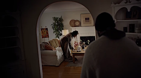 Indoor domestic scene featuring two adults in a living room; one standing, tending to a coffee table, the other lying on a couch.