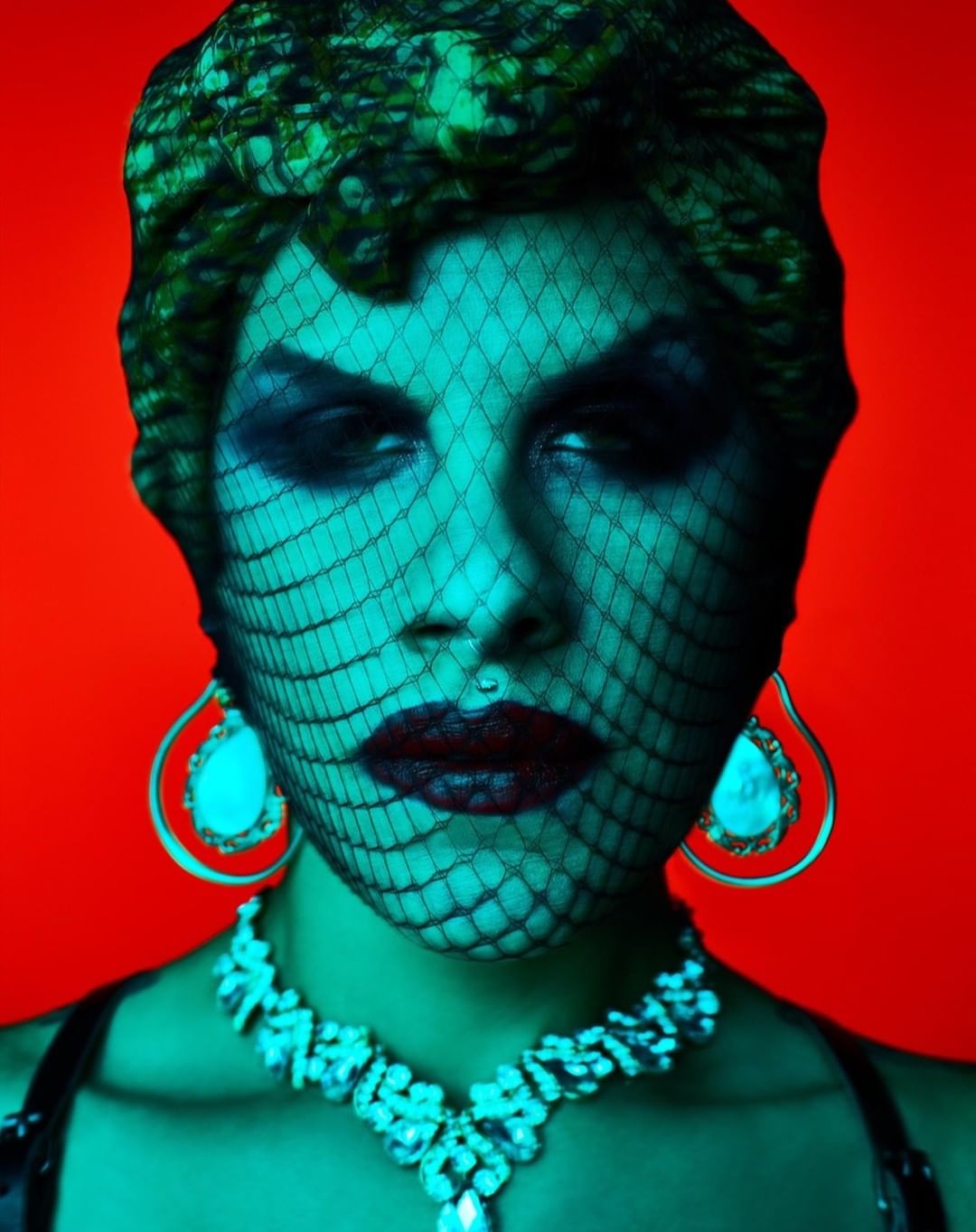 Portrait of a woman with bold makeup, a net veil, and ornate jewelry against a vibrant red backdrop, creating a striking and theatrical image.