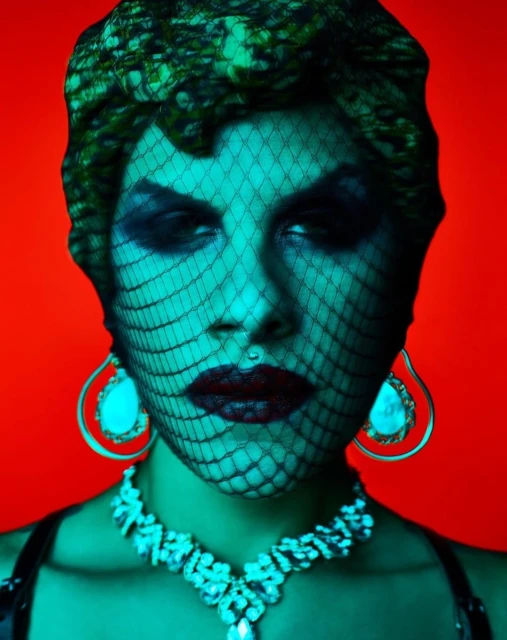 Portrait of a woman with bold makeup, a net veil, and ornate jewelry against a vibrant red backdrop, creating a striking and theatrical image.