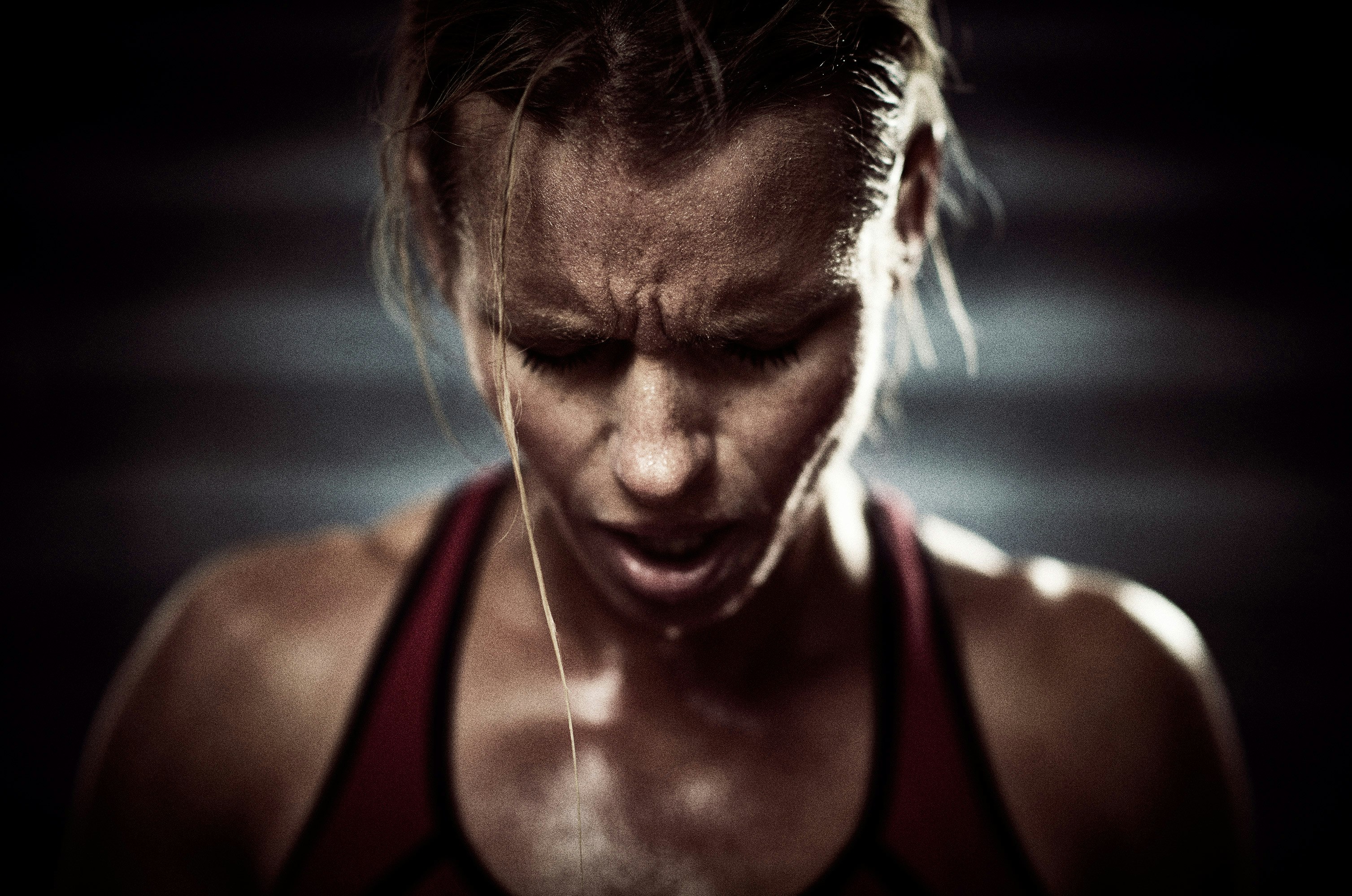 Close-up of a sweaty adult female athlete with an intense expression, wearing a sports bra, in a dimly lit indoor fitness setting.