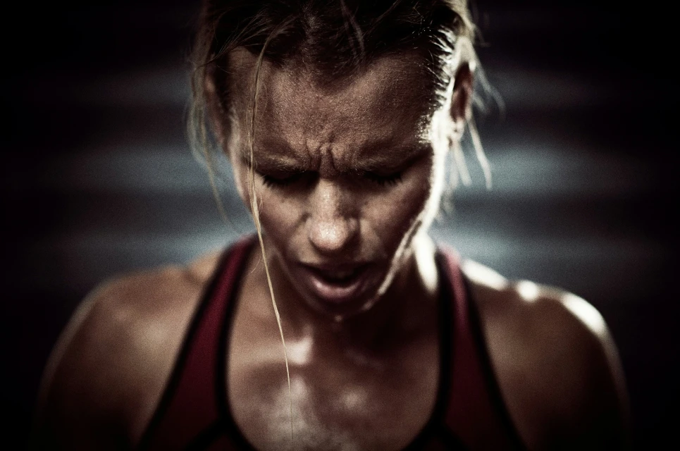 Close-up of a sweaty adult female athlete with an intense expression, wearing a sports bra, in a dimly lit indoor fitness setting.