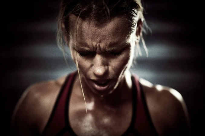 Close-up of a sweaty adult female athlete with an intense expression, wearing a sports bra, in a dimly lit indoor fitness setting.