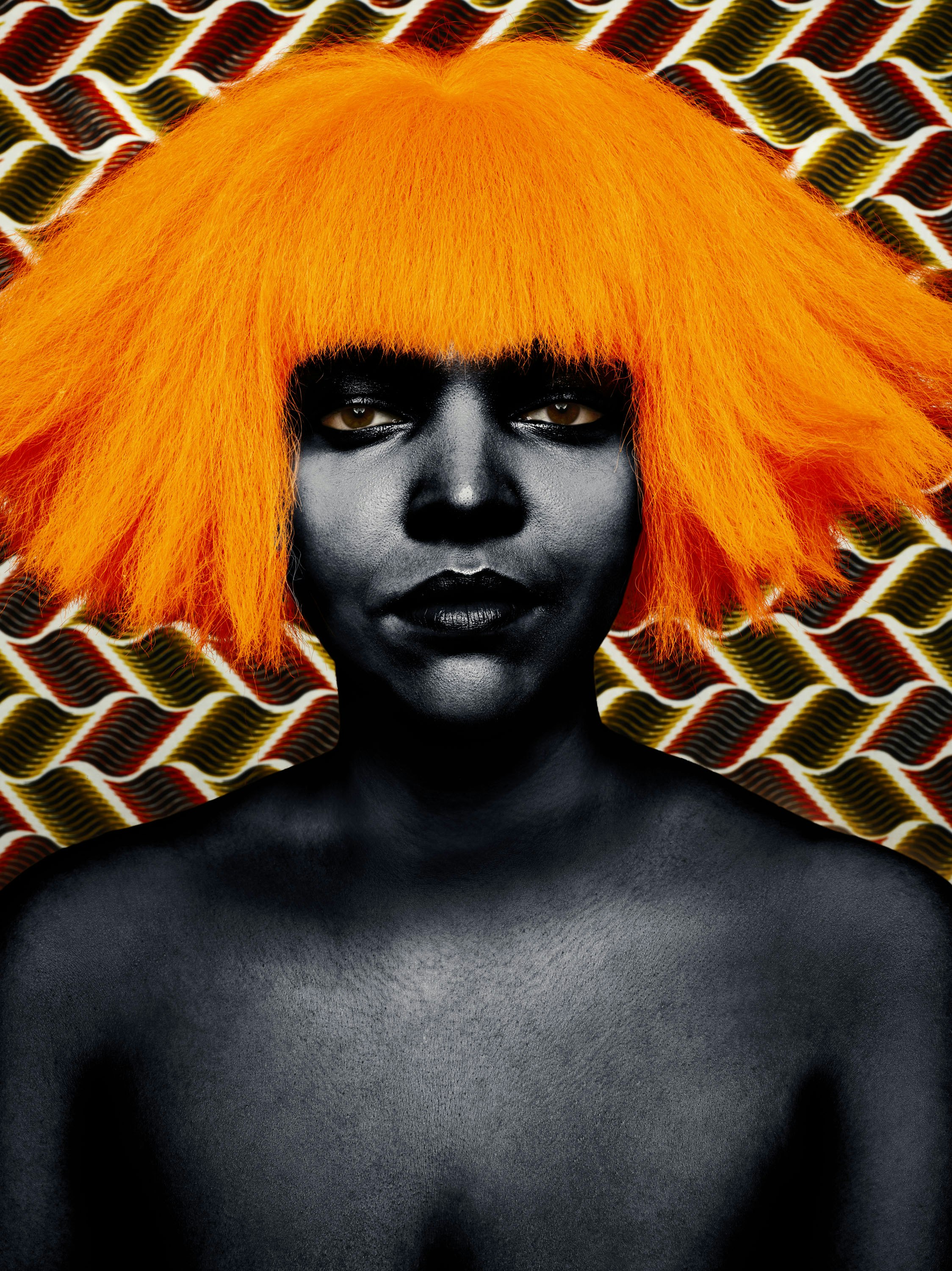 Dark-skinned individual with vibrant orange wig against a geometric patterned backdrop. Striking contrast between skin tone and wig color.