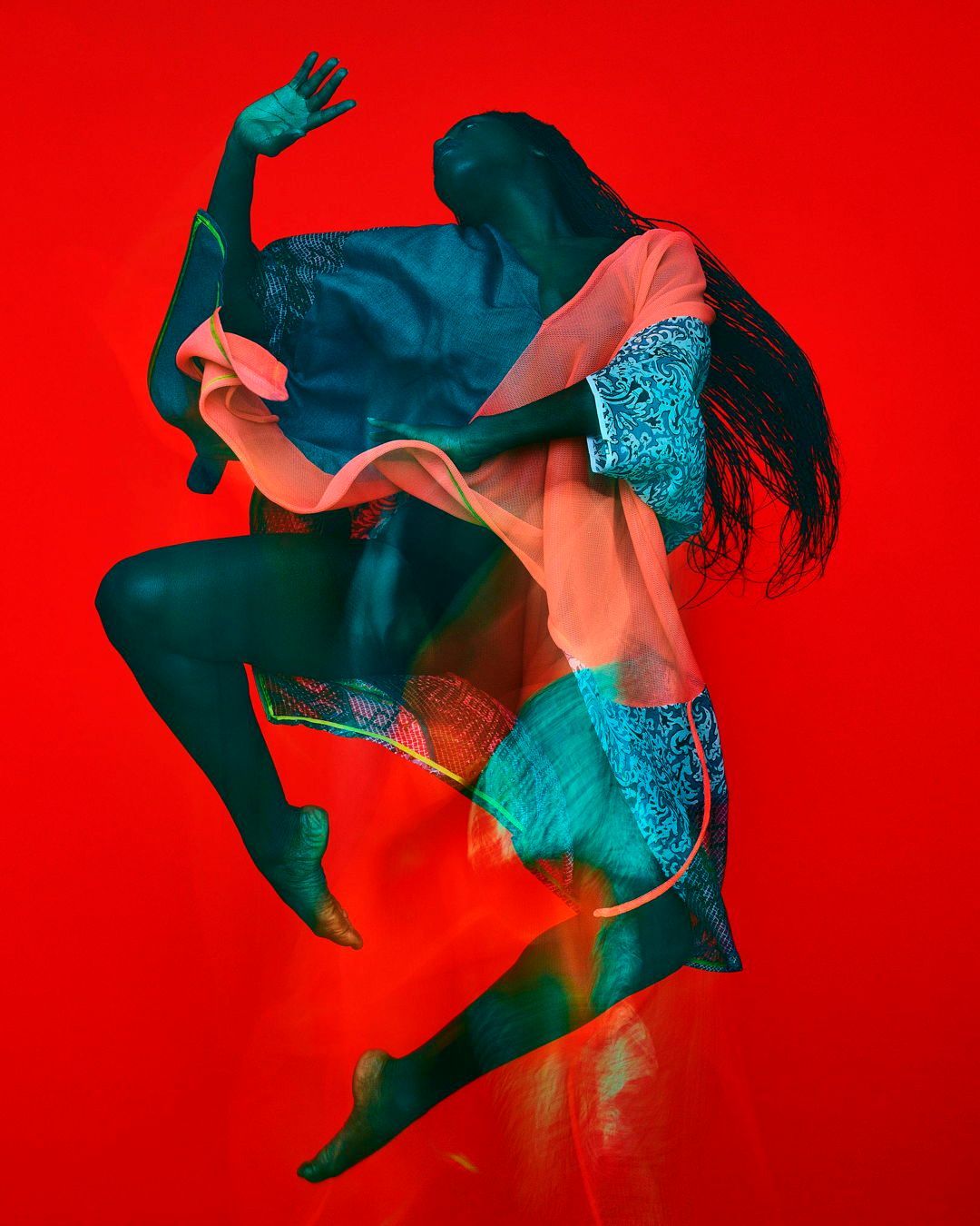 Fashion shot of a woman in motion with dark skin and flowing black hair. She wears a peach colored top, on a bright red backdrop.