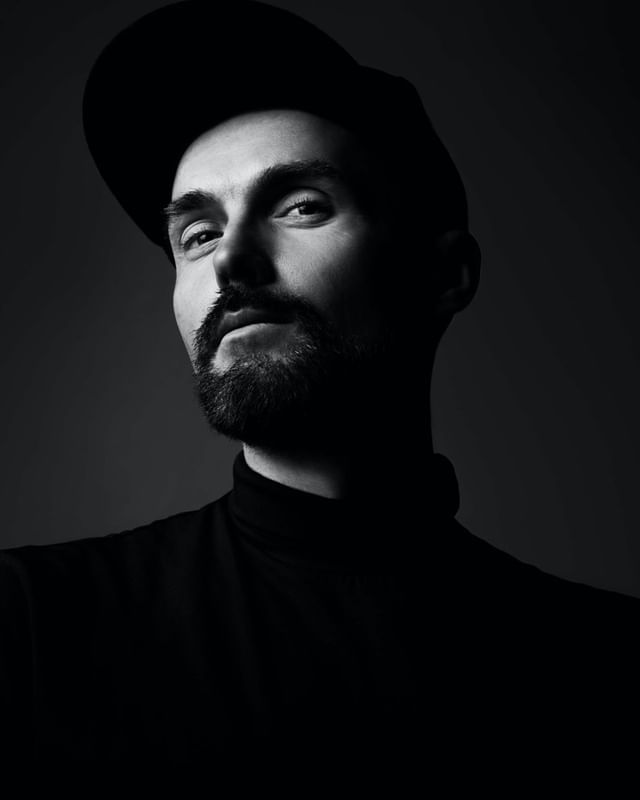 Monochromatic portrait of a man with a beard wearing a cap and turtleneck, looking at the camera in a photo studio with dark gray background.