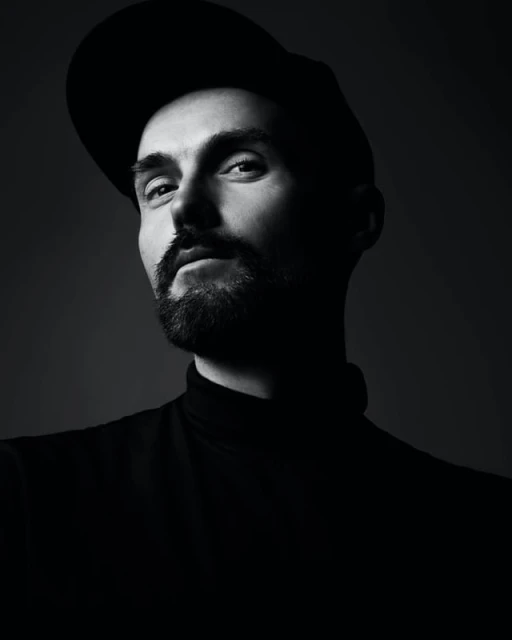 Monochromatic portrait of a man with a beard wearing a cap and turtleneck, looking at the camera in a photo studio with dark gray background.