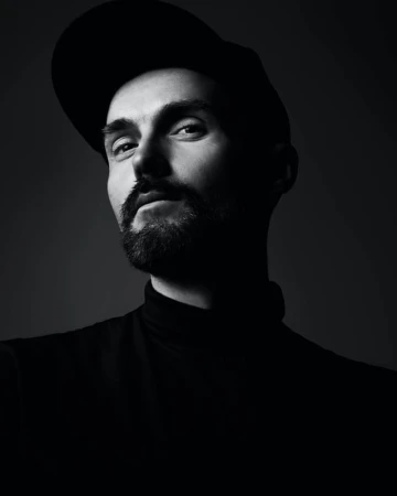 Monochromatic portrait of a man with a beard wearing a cap and turtleneck, looking at the camera in a photo studio with dark gray background.