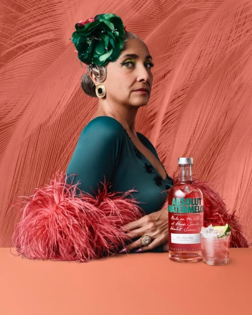 Elegant woman with a green floral headpiece and a feathered boa posing with an Absolut Watermelon bottle and cocktail against a coral backdrop.