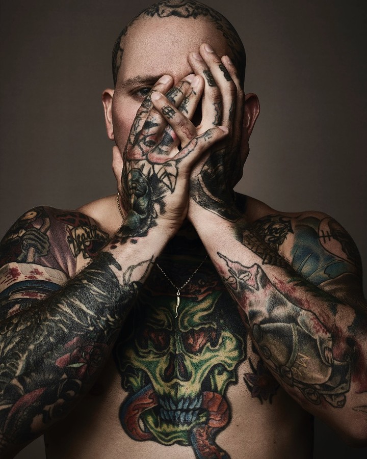 Close-up portrait of a bare-chested adult male covered in tattoos, concealing his face with tattooed hands against a neutral backdrop.