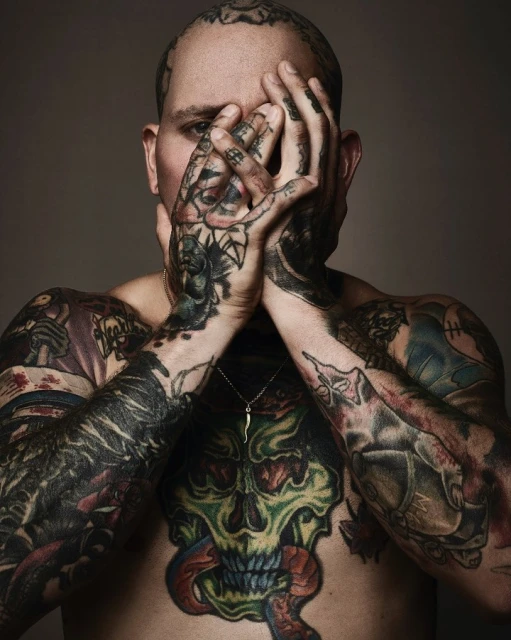 Close-up portrait of a bare-chested adult male covered in tattoos, concealing his face with tattooed hands against a neutral backdrop.