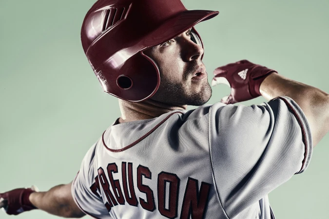 Male baseball player in uniform and helmet looking up.  Baseball player ready to swing, posing against a light green backdrop.