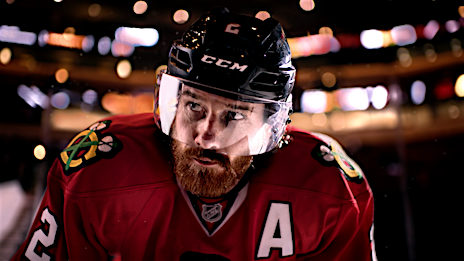 Close-up of a male ice hockey player in team jersey and helmet, looking up during a game; arena lights are blurred in the background.