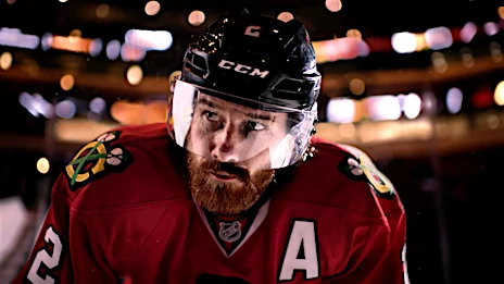 Close-up of a male ice hockey player in team jersey and helmet, looking up during a game; arena lights are blurred in the background.