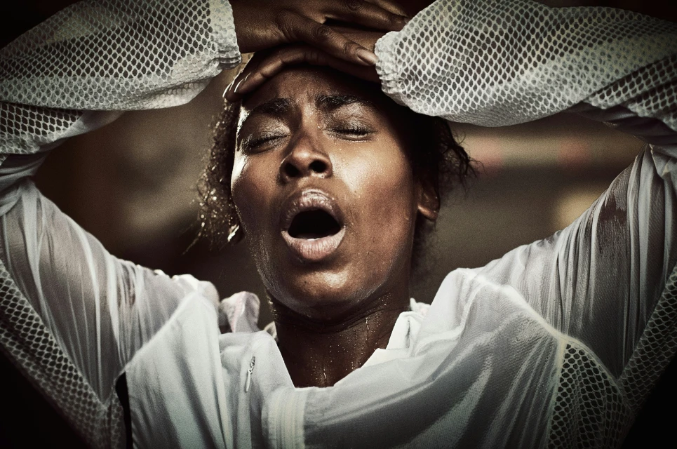 Close-up portrait of an African American woman with her eyes closed and mouth open, hands on her head, wearing a white net jacket.
