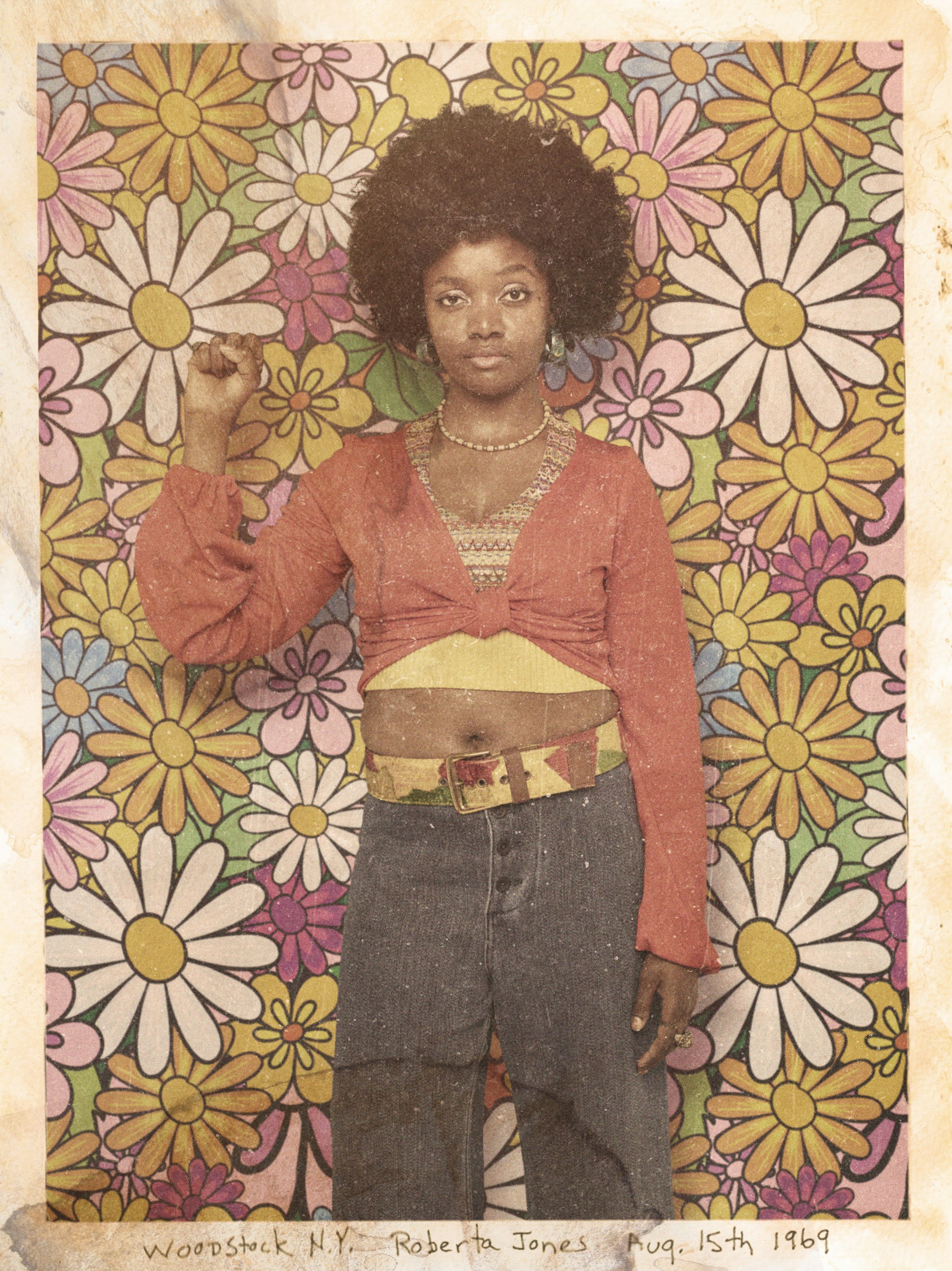 Portrait of a woman with an afro hairstyle, orange top, and jeans, with her fist raised against a floral backdrop.