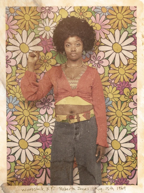 Portrait of a woman with an afro hairstyle, orange top, and jeans, with her fist raised against a floral backdrop.