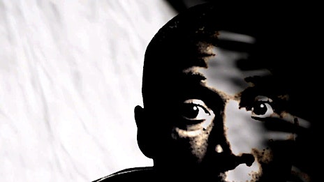 Close-up portrait of a young Black person in dark lighting against a white background, highlighting their eyes and facial features.