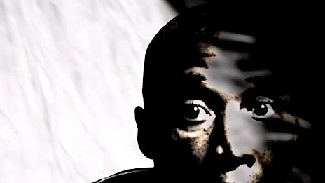 Close-up portrait of a young Black person in dark lighting against a white background, highlighting their eyes and facial features.