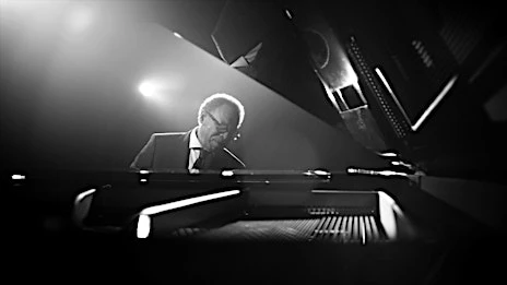 Monochrome shot of a mature man in a suit playing a grand piano with dramatic spotlighting in an indoor performance setting.