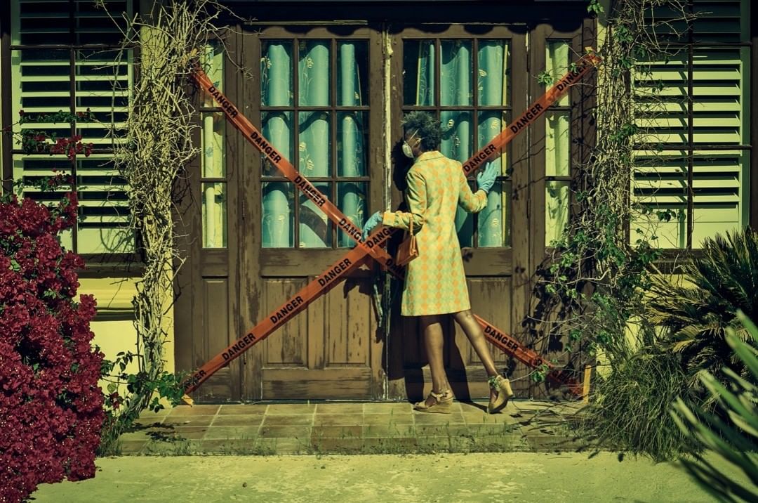 Woman wearing a yellow patterned coat and face mask applying caution tape on the front door with flowering plants nearby.