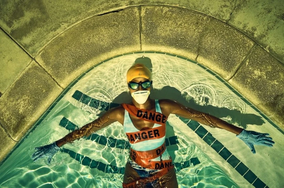 Overhead shot of a woman floating in a swimming pool, wearing a swim cap, goggles, gloves, and a mask with 'DANGER' tape across her.
