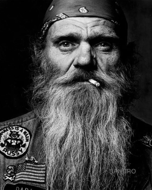 Monochrome close-up portrait of an adult male with a long beard, wearing a bandana and jacket with patches, smoking a cigarette.